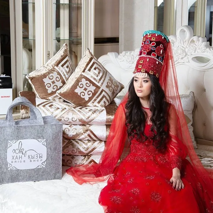 Model in a red traditional Kazakh bridal dress beside Xan Kyzy bedding and a branded bag
